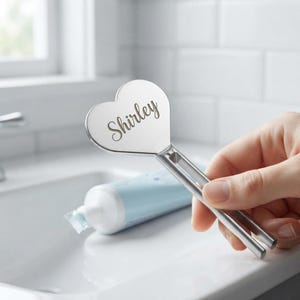 May include: A silver-colored metal tube squeezer featuring a heart-shaped top with the name Shirley engraved in a cursive script. The tool has a long, slotted handle designed to slide over the end of a tube to dispense its contents.