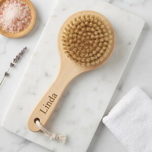 May include: A round wooden bath brush with natural tan bristles and a long handle. The handle features the name Linda engraved in a dark font and includes a small hole at the end with a short, off-white cotton rope loop attached for hanging.
