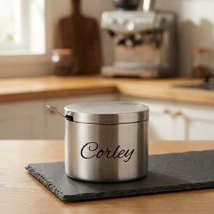 May include: A cylindrical stainless steel canister with a matching lid and a small metal spoon inserted through a side notch. The exterior features the name Corley in a black cursive script font, providing a clear label for the container.