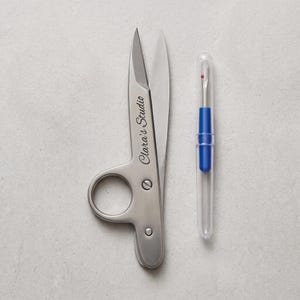 May include: A pair of silver-toned metal thread snips with the text Clara's Studio engraved on the blade, positioned next to a blue seam ripper with a clear plastic protective cap. Both tools are designed for sewing and textile craft tasks.