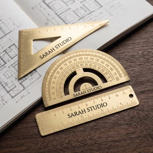 May include: A set of three brass drafting tools including a triangular ruler, a semi-circular protractor, and a straight ruler, each engraved with the text SARAH STUDIO. The tools feature clear measurement markings in both inches and centimeters.