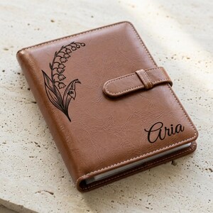 May include: A brown leather-like journal with a textured finish, featuring a black engraved lily of the valley floral design on the left side and the name Aria in a cursive script on the right, secured by a matching horizontal strap closure.