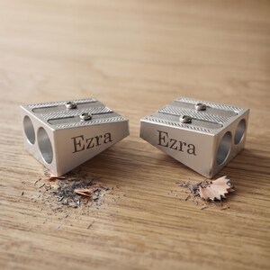 May include: Two silver-colored metal pencil sharpeners with a wedge shape, each featuring two circular openings for different pencil sizes and the name Ezra engraved on the side. The sharpeners have visible metal blades and screws.