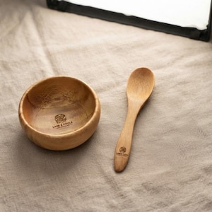 May include: A small, round, light brown wooden bowl and a matching wooden spoon. The interior of the bowl and the handle of the spoon both feature a dark brown circular logo with the text "LASE & POOLS" printed in a clear, sans-serif font.