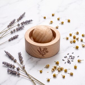 May include: A round wooden mortar and pestle set featuring a light brown natural wood grain. The exterior of the mortar is engraved with a small floral bouquet illustration. The set is surrounded by dried lavender sprigs and chamomile flowers.