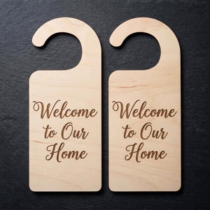 May include: Two light-colored wooden door hangers with a smooth finish. Each hanger features a curved hook at the top and the engraved text Welcome to Our Home in a dark brown cursive script centered on the rectangular body of the wood piece.