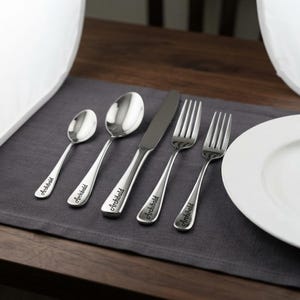 May include: A five-piece stainless steel flatware set consisting of a small spoon, a large spoon, a dinner knife, a dinner fork, and a salad fork. Each piece features a polished silver finish and the name Archibald engraved in a cursive script.