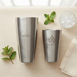 May include: Two brushed stainless steel tumblers of different sizes. The larger tumbler is engraved with the name Kathleen. The smaller tumbler features a floral emblem above the name Ambrella. Fresh green mint leaves and a bowl of ice cubes.