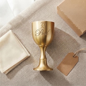 May include: A metallic gold-toned goblet featuring a smooth, brushed finish and a pedestal base. The exterior is engraved with the letter P in a decorative font above the cursive script name Poul. The goblet has a wide bowl and a slender stem.