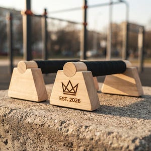 May include: A pair of light-colored wooden push-up bars with black grip tape wrapped around the center handles. The front wooden base features a laser-engraved crown icon above the text EST. 2026, showcasing a sturdy, functional design.