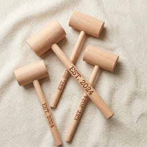 May include: Four light-colored wooden mallets arranged in a cluster. Each mallet features a cylindrical head and a smooth handle. Three of the handles are engraved with the text EST. 2024, while one handle shows the partial text EST. 202.