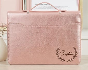 May include: A rose gold metallic portfolio case with a top handle and a horizontal flap. The front features a dark brown engraved design of a laurel wreath surrounding the name Sophia in a cursive script font, centered on the lower right side.