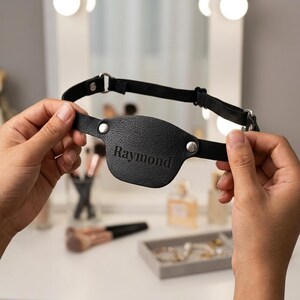 May include: A black leather eye patch featuring the name Raymond embossed on the front. The patch is attached to a thin, adjustable black elastic strap with silver-toned metal hardware, including circular rivets and a sliding buckle for sizing.