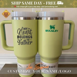 May include: Two green stainless steel travel mugs with lids. The mug on the left has the text "My Heart Belongs to Father" on it. The mug on the right has the Stanley logo and the text "Quencher H2.0 Tumbler".