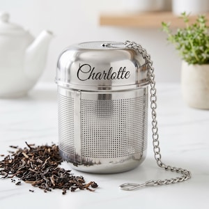 May include: A cylindrical stainless steel tea infuser with a fine mesh body and a solid top lid. The name Charlotte is engraved in a cursive script on the side of the lid. A metal chain with a small hook is attached to the top of the infuser.