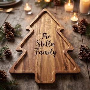 May include: A Christmas tree-shaped wooden serving board with a natural wood grain finish. The center of the board features the engraved text The Stella Family in a dark script font. The board has a raised edge and a smooth, polished surface.