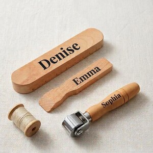 May include: A wooden sewing tool set featuring a long rectangular block engraved with the name Denise, a smaller rectangular block engraved with Emma, a wooden roller tool engraved with Sophia, and a small wooden spool with light beige thread.