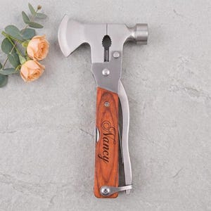 May include: A multi-tool hammer featuring a silver metal head with an integrated axe blade and a wooden handle engraved with the name Nancy in a cursive script. The tool includes a nail puller and a hammer face, with a smooth, polished finish.