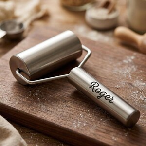 May include: A stainless steel rolling pin featuring a cylindrical barrel and a handle engraved with the name Roger in a cursive script. The metal tool has a smooth, polished finish and is connected by a sturdy wire frame structure.