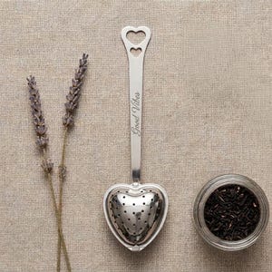 May include: A silver-toned metal tea infuser with a heart-shaped perforated head and a long handle featuring two heart cutouts at the top. The handle is engraved with the text Good Vibes. Two sprigs of dried lavender and a glass jar of loose tea.