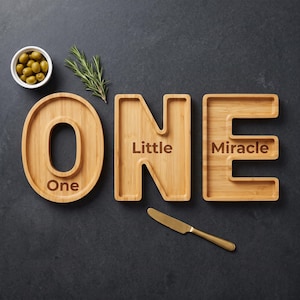May include: Three bamboo serving trays shaped like the letters O, N, and E. The O tray is engraved with the word One, the N tray with Little, and the E tray with Miracle. A small white bowl filled with green olives, a sprig of rosemary, and a gold knife.