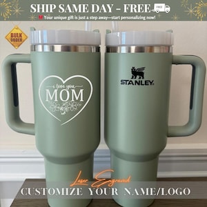 May include: Two green stainless steel travel mugs with lids. One mug has the words "I love you MOM" engraved on it. The other mug has the Stanley logo engraved on it. Both mugs have a handle.