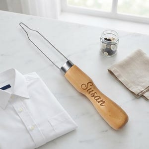 May include: A button hook tool featuring a long, thin metal wire loop attached to a light-colored wooden handle. The handle is engraved with the name Susan in a cursive script. The tool is positioned next to a white shirt and a glass jar of buttons.