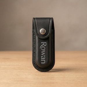May include: A black leather pouch with a rounded bottom and a flap closure secured by a silver-toned metal snap button. The front of the pouch features the name Rowan engraved in a vertical orientation with a clean, serif-style font.