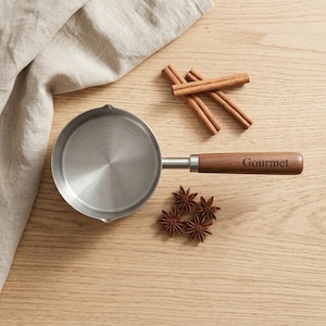 May include: A small stainless steel saucepan with a wooden handle featuring the word Gourmet engraved on it. The pan is accompanied by three star anise pods and three cinnamon sticks arranged nearby on a light-colored wooden surface.
