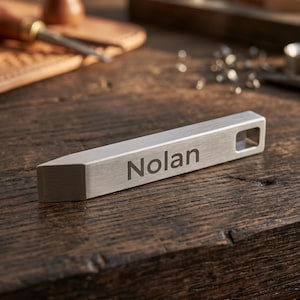 May include: A rectangular silver-toned metal bar with a brushed finish, featuring the name Nolan engraved in a dark sans-serif font on the front face. The object has a beveled end and a small square hole at the opposite end, presenting a clean design.