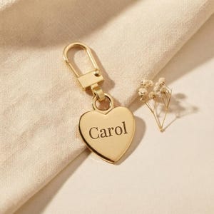 May include: A gold-toned metal keychain featuring a heart-shaped pendant with the name Carol engraved in a serif font. The pendant is attached to a rectangular lobster clasp mechanism, creating a polished and functional accessory for keys.