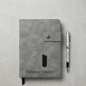 May include: A gray notebook with a textured cover featuring a diagonal pocket, a snap button closure, and the name Eleanor Vance printed at the bottom. A silver metal pen with the name E. Vance engraved on the barrel is positioned next to the notebook.