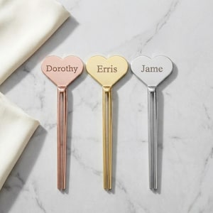 May include: Three metal heart-shaped food picks with two-pronged stems. The left pick is rose gold with the name Dorothy, the center pick is gold with the name Erris, and the right pick is silver with the name Jame engraved on the heart faces.
