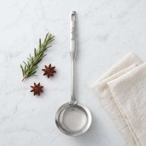 May include: A silver-toned metal skimmer spoon with a circular mesh head and a long handle featuring the engraved name Sophia. The utensil has a smooth finish and a rectangular hanging hole at the top of the handle, designed for kitchen use.