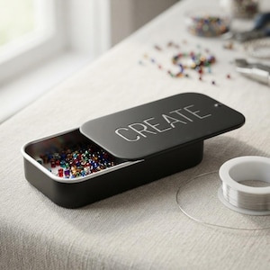May include: A rectangular black metal storage tin with a sliding lid featuring the word CREATE embossed in white. The tin is partially open, revealing a collection of small, multicolored glass beads stored inside the container.