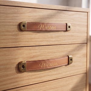 May include: A light-colored wooden drawer unit featuring two horizontal tan leather drawer pulls. Each leather strap is secured with antique brass-toned metal hardware at both ends and features the name Weston embossed in a cursive script.