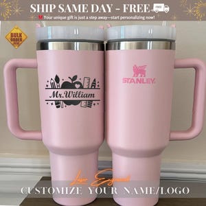 May include: Two pink stainless steel travel mugs with lids. One mug has a black design of a teacher's apple with school supplies and the name "Mr. William". The other mug has a black logo of a bird with the word "STANLEY" underneath.