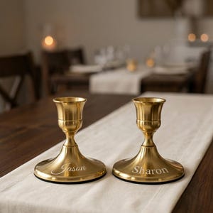 May include: Two matching gold-toned metal candle holders with a smooth, polished finish. Each holder features a wide, circular base engraved with a name in a distinct font: one reads Jason in script, and the other reads Sharon in a serif typeface.