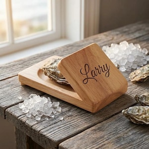 May include: A light brown wooden oyster serving tray with a hinged lid engraved with the name Larry in a cursive script. The tray features a circular indentation holding a single oyster shell, accompanied by scattered clear ice crystals.