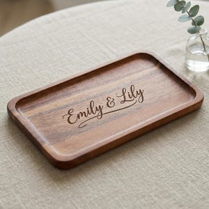 May include: A rectangular wooden tray with rounded corners and a natural brown wood grain finish. The center of the tray features the engraved text "Emily & Lily" in a cursive script font. The tray has a smooth, shallow surface design.
