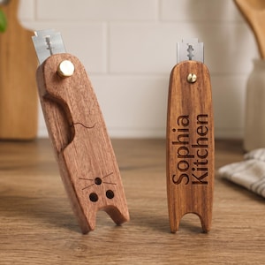 May include: Two wooden bread lames with metal blades secured by brass knobs. One handle is shaped like a cat with engraved whiskers and eyes, while the other is a straight handle engraved with the text Sophia Kitchen in a dark sans-serif font.