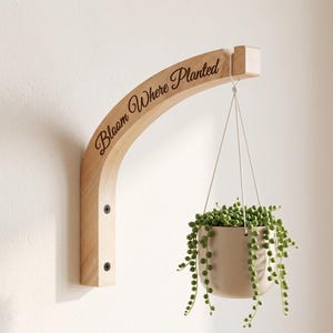 May include: A light-colored wooden wall-mounted plant hanger with a curved arm featuring the engraved text "Bloom Where Planted." A white ceramic pot hangs from the arm by thin white cords, containing a trailing green string of pearls plant.