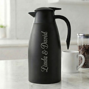 May include: A matte black thermal carafe with a curved handle and a secure lid. The side of the carafe features the engraved text Linda & David in a cursive script font. The carafe is designed for holding hot or cold beverages for serving.