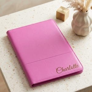 May include: A bright pink rectangular notebook with a smooth, solid-colored cover. The name Charlotte is embossed in a dark, elegant script font on the lower right corner of the front cover. The notebook features a horizontal seam across the lower section.