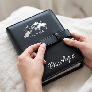 May include: A black leather-like journal featuring a silver-colored graphic of clouds and a sun on the top cover. The name Penelope is printed in a white cursive script on the bottom right. A matching black strap closure secures the front cover.