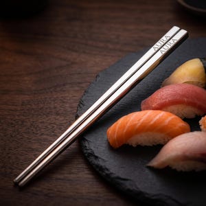 May include: A pair of sleek, silver-toned metal chopsticks with a square profile. The word AURA is engraved in a clean, sans-serif font near the top end of each chopstick. The chopsticks are positioned diagonally across a dark, textured slate plate.