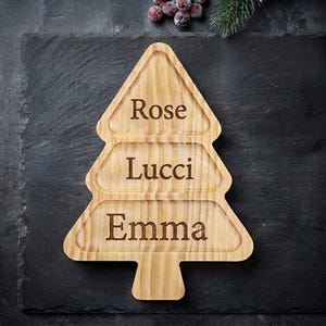 May include: A light brown wooden serving tray shaped like a Christmas tree with three horizontal sections. The names Rose, Lucci, and Emma are engraved in dark lettering within each of the three sections of the wooden tree-shaped tray.
