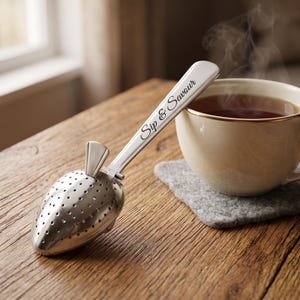 May include: A silver-toned metal tea infuser shaped like a strawberry with a perforated body and a long handle. The handle is engraved with the text "Sip & Savour" in a cursive script. The infuser is positioned next to a steaming cup of tea.