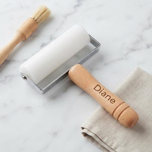 May include: A white rubber brayer with a silver metal frame and a light wood handle engraved with the name Diane, accompanied by a wooden-handled brush with light-colored bristles and a folded beige linen cloth on a white marble surface.