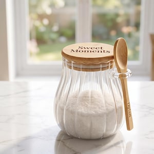 May include: A clear, ribbed glass jar filled with white sugar, featuring a round bamboo lid engraved with the text "Sweet Moments." A matching bamboo spoon is held in a glass loop on the side of the jar, with the name "Dylan" engraved on the handle.